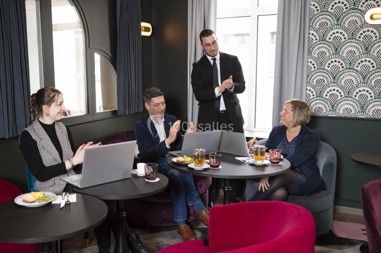 Quatre personnes discutent et applaudissent autour de tables avec ordinateurs et boissons dans un salon moderne.