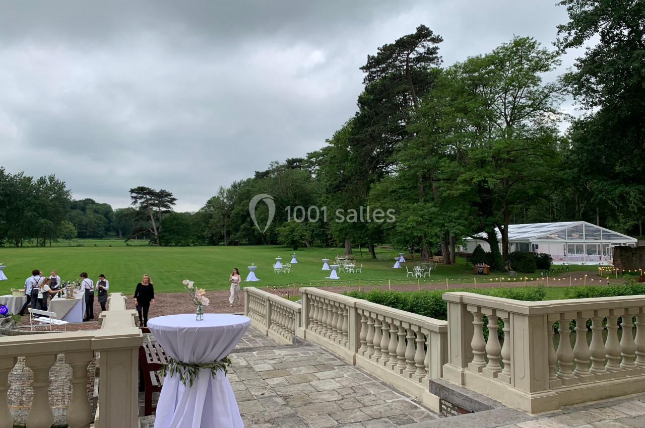 Terrasse en pierre surplombant un grand jardin verdoyant avec des tables dressées et une tente blanche en arrière-plan.