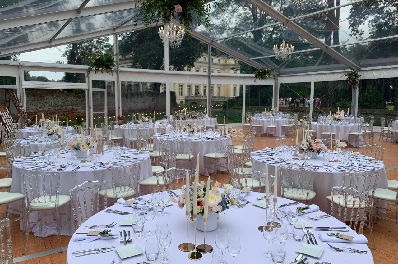 Salle de réception sous une tente transparente, tables rondes dressées avec nappes blanches et décorations florales.