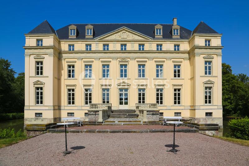 Façade d'un château classique à trois étages, entouré de verdure, sous un ciel bleu dégagé.