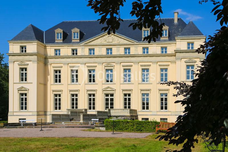 Façade d'un château de style classique avec toit en ardoise, entouré de végétation et éclairé par le soleil.