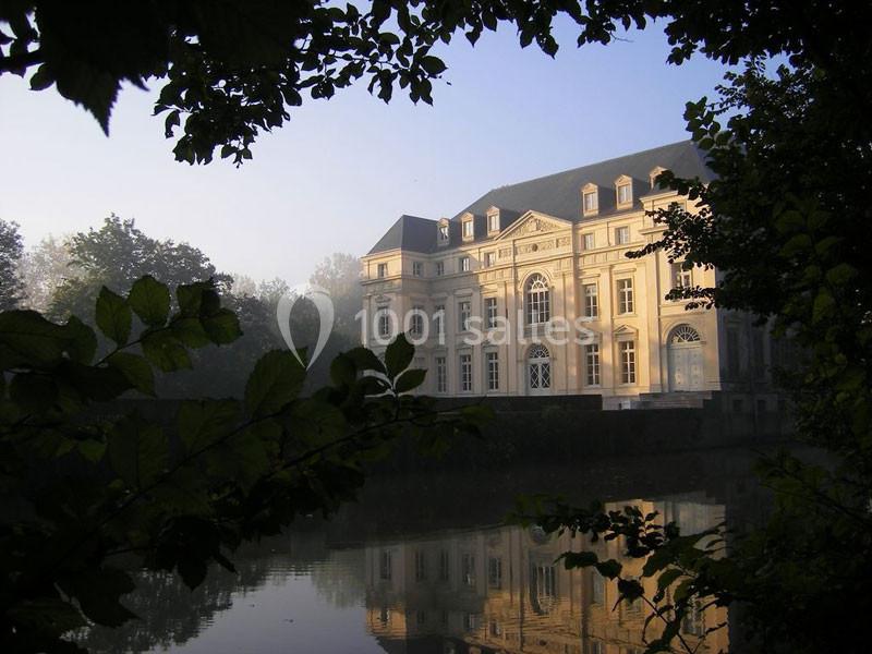 Façade d'un château éclairé par le soleil, entouré de végétation et reflété dans l'eau calme d'un étang.