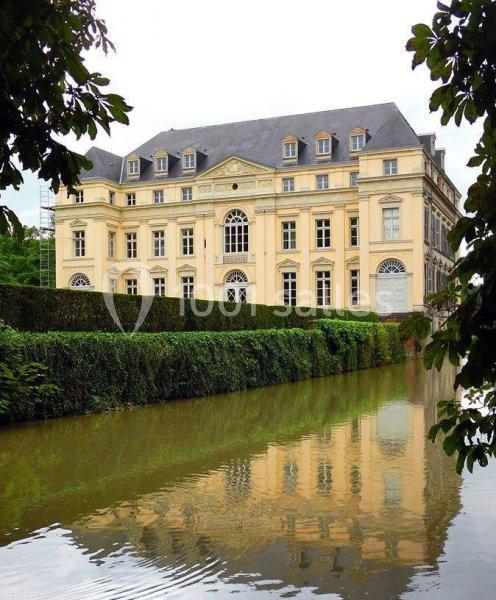 Façade d'un bâtiment historique beige à plusieurs étages, bordé par une haie et un canal reflétant son image.