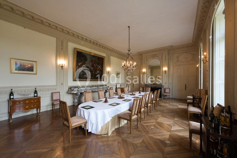 Salle élégante avec parquet en bois, table dressée, chaises alignées, lustre central et tableaux accrochés aux murs.