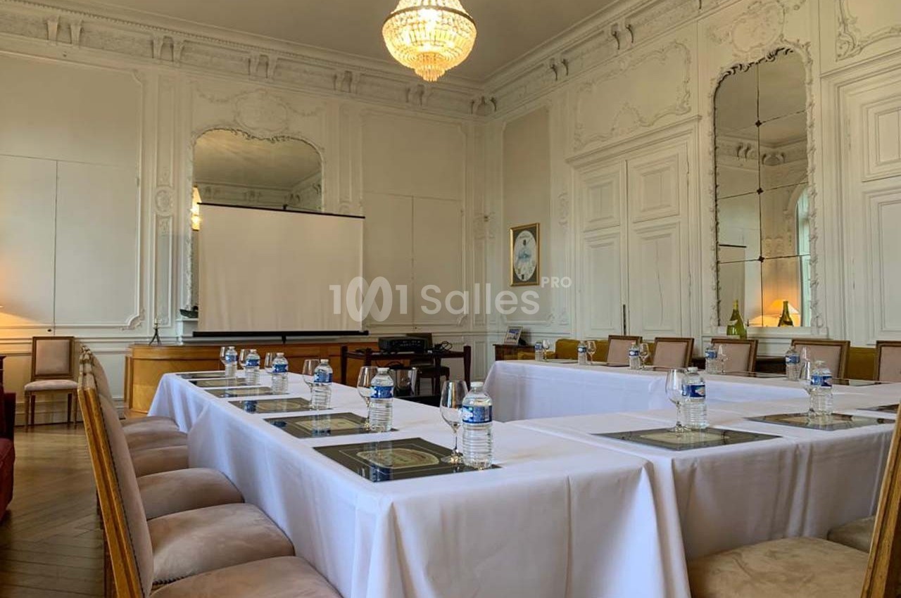 Salle de réunion élégante avec tables disposées en U, chaises rembourrées, écran de projection et décor classique.