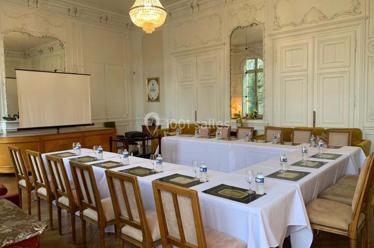 Salle de réunion élégante avec tables disposées en U, chaises en bois, écran de projection et lustre au plafond.
