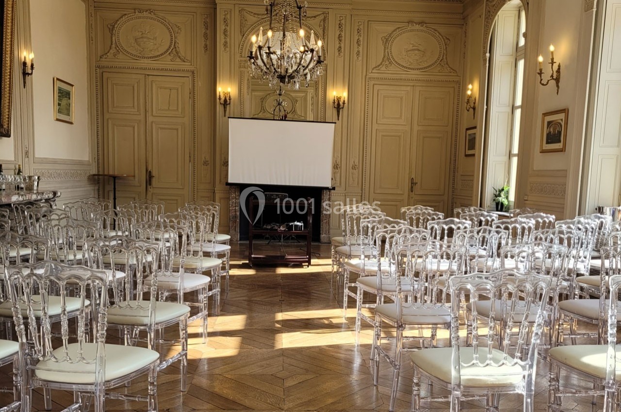 Salle élégante avec parquet, chaises transparentes alignées et un écran de projection devant une cheminée.