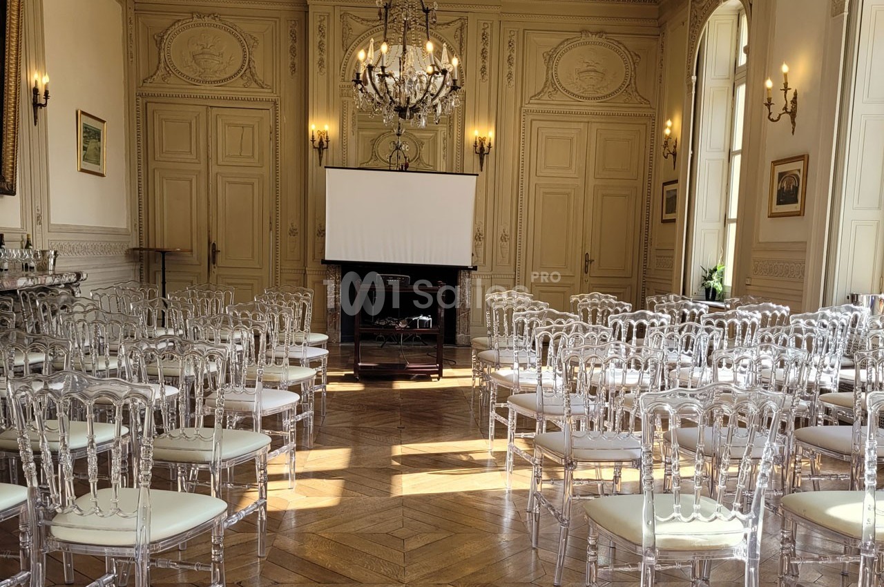 Salle élégante avec parquet, chaises transparentes alignées et un écran de projection devant une cheminée.