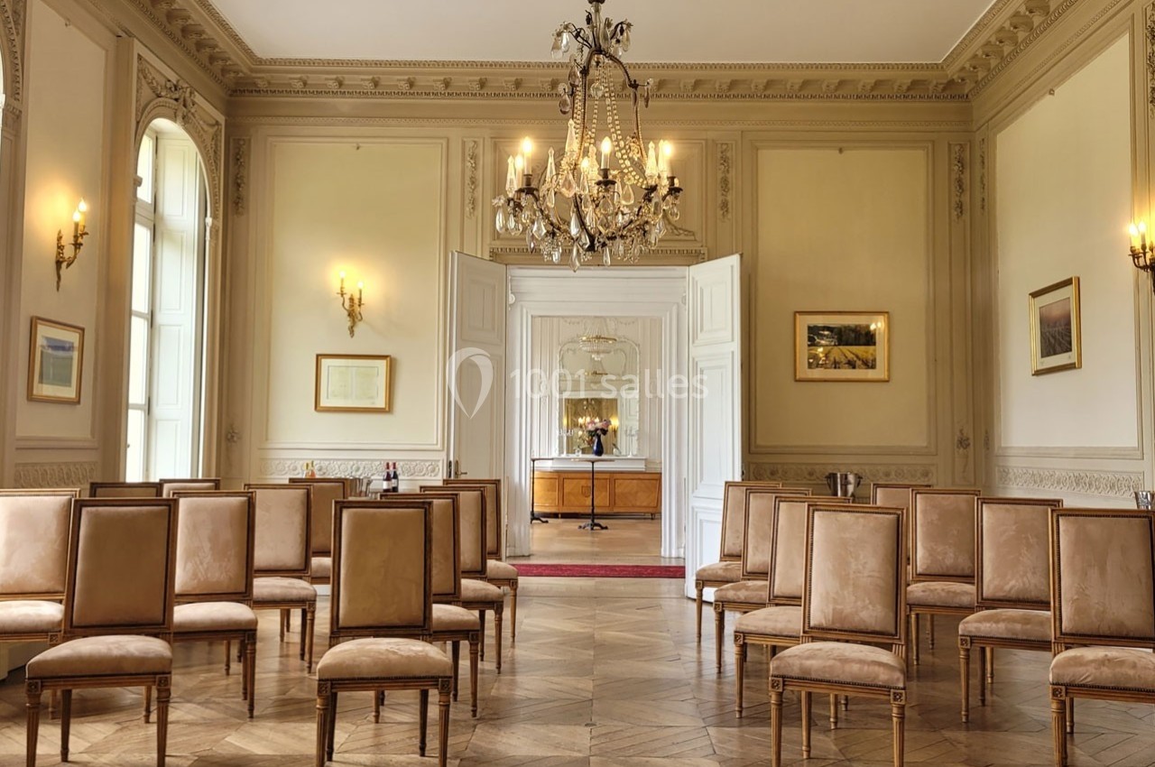 Salle lumineuse avec parquet en bois, chaises alignées, murs ornés de moulures et lustre central.