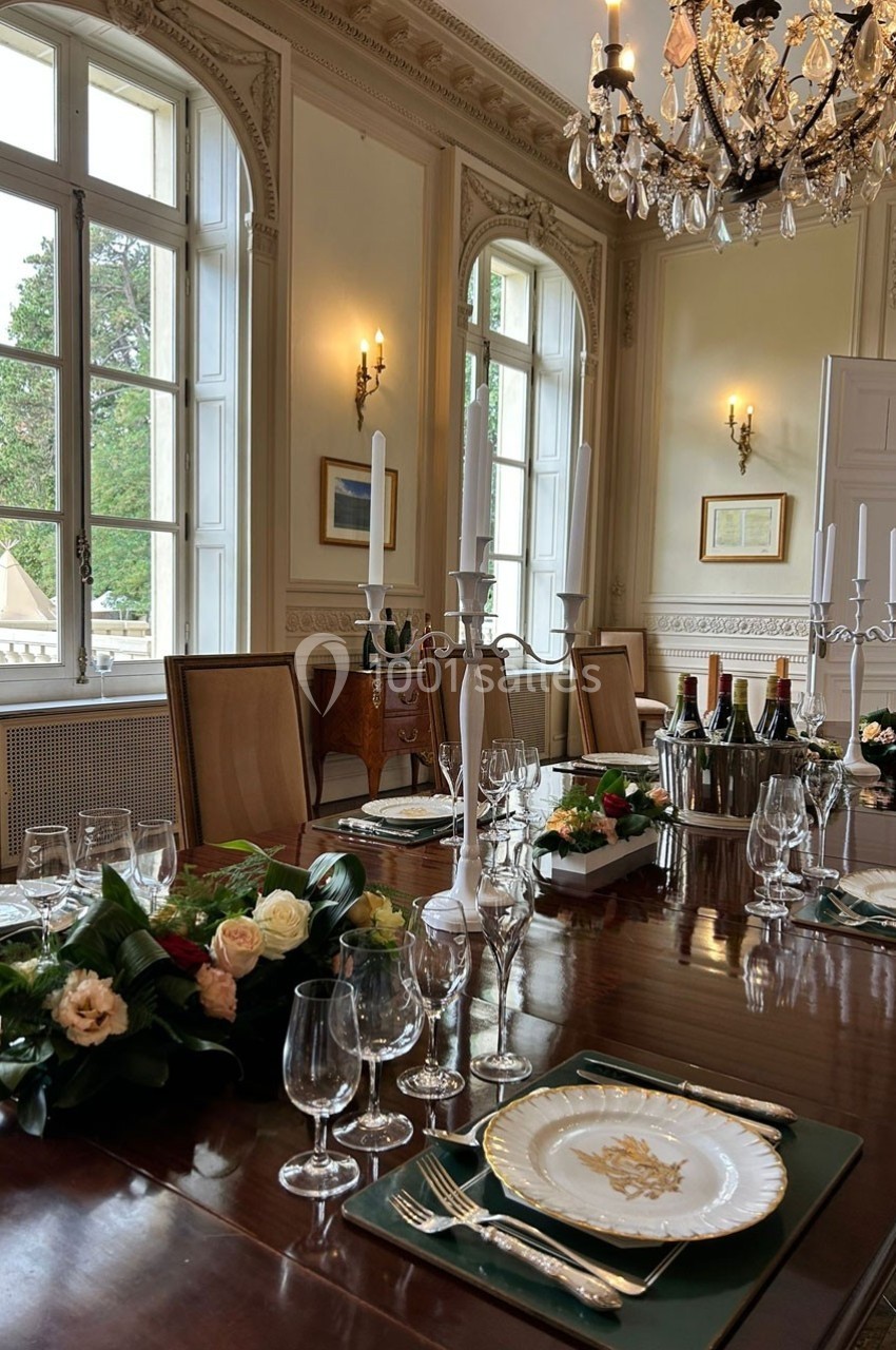 Table élégamment dressée avec vaisselle raffinée, chandeliers et décorations florales dans une salle lumineuse et classique.