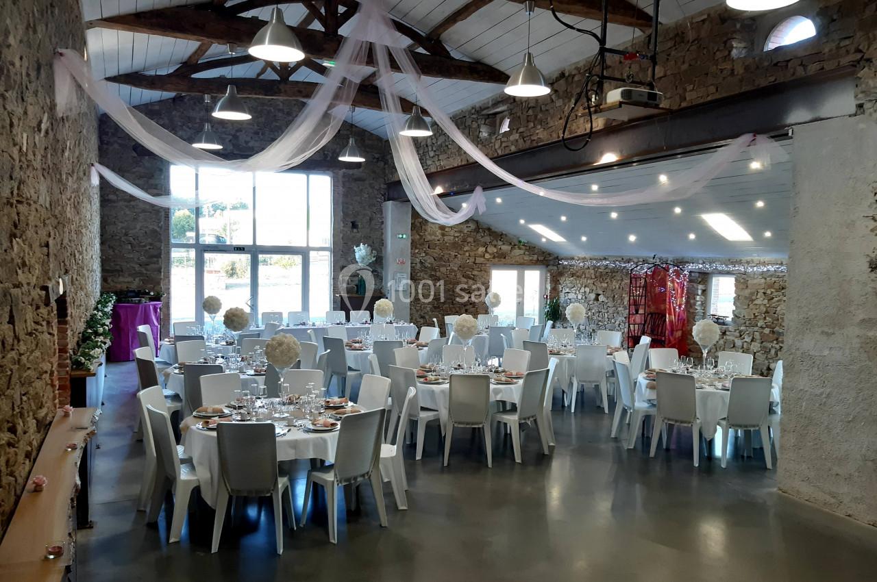 Salle de réception en pierre avec tables dressées pour un événement, décorée de voilages suspendus et éclairée naturellement.