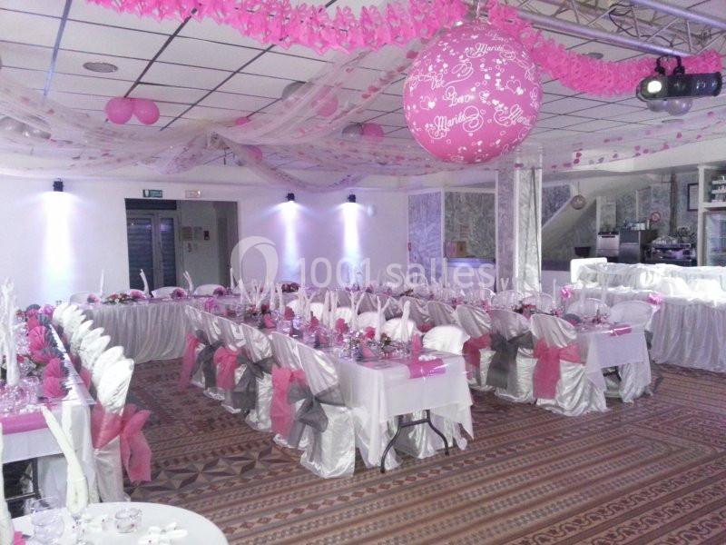 Salle de réception décorée en rose et blanc avec tables dressées, chaises ornées de nœuds et ballons suspendus.