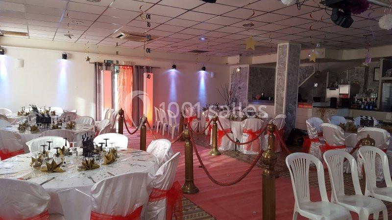Salle de réception décorée avec des tables rondes, nappes blanches, chaises et rubans rouges, séparée par des cordons dorés.