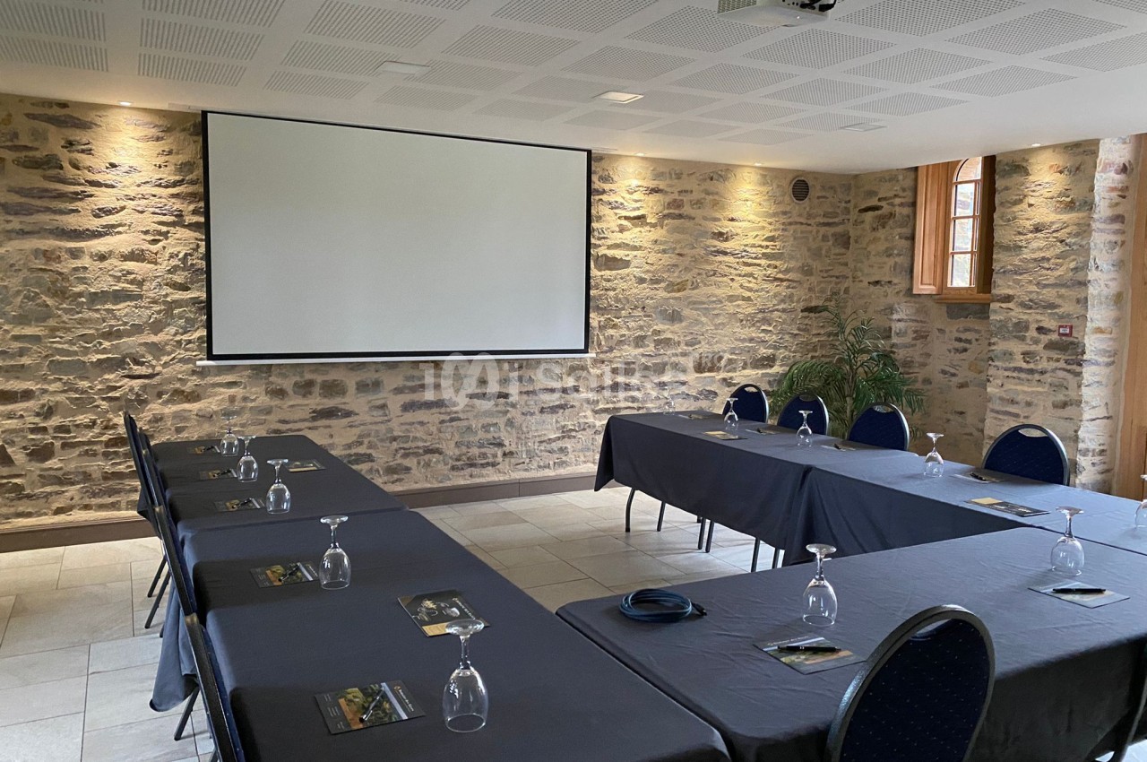 Salle de réunion avec murs en pierre, tables disposées en U, verres et documents posés, écran de projection au fond.