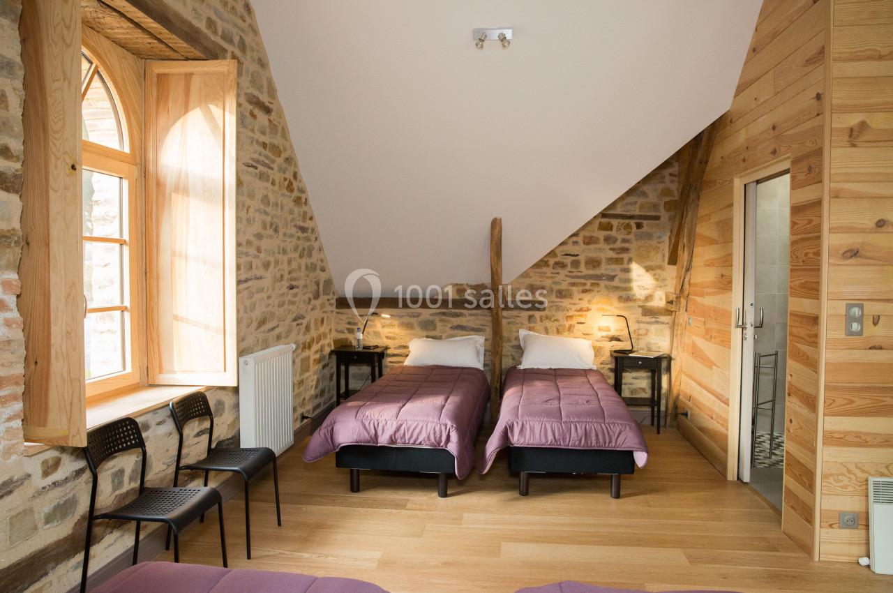 Chambre avec deux lits simples, murs en pierre et bois, fenêtre à arc, parquet clair et salle de bain attenante.