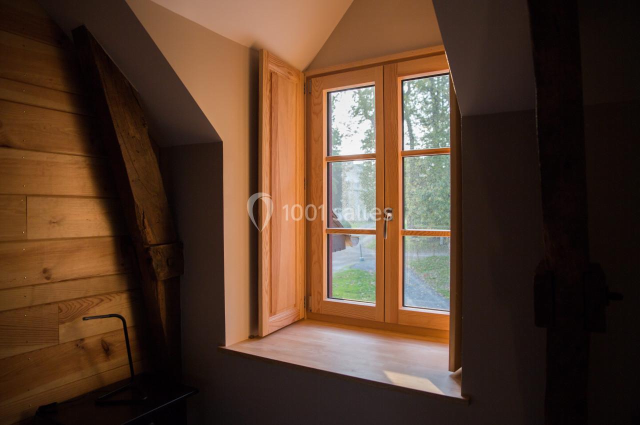 Fenêtre en bois ouverte sur un paysage extérieur verdoyant, éclairant une pièce aux murs en bois et poutres apparentes.