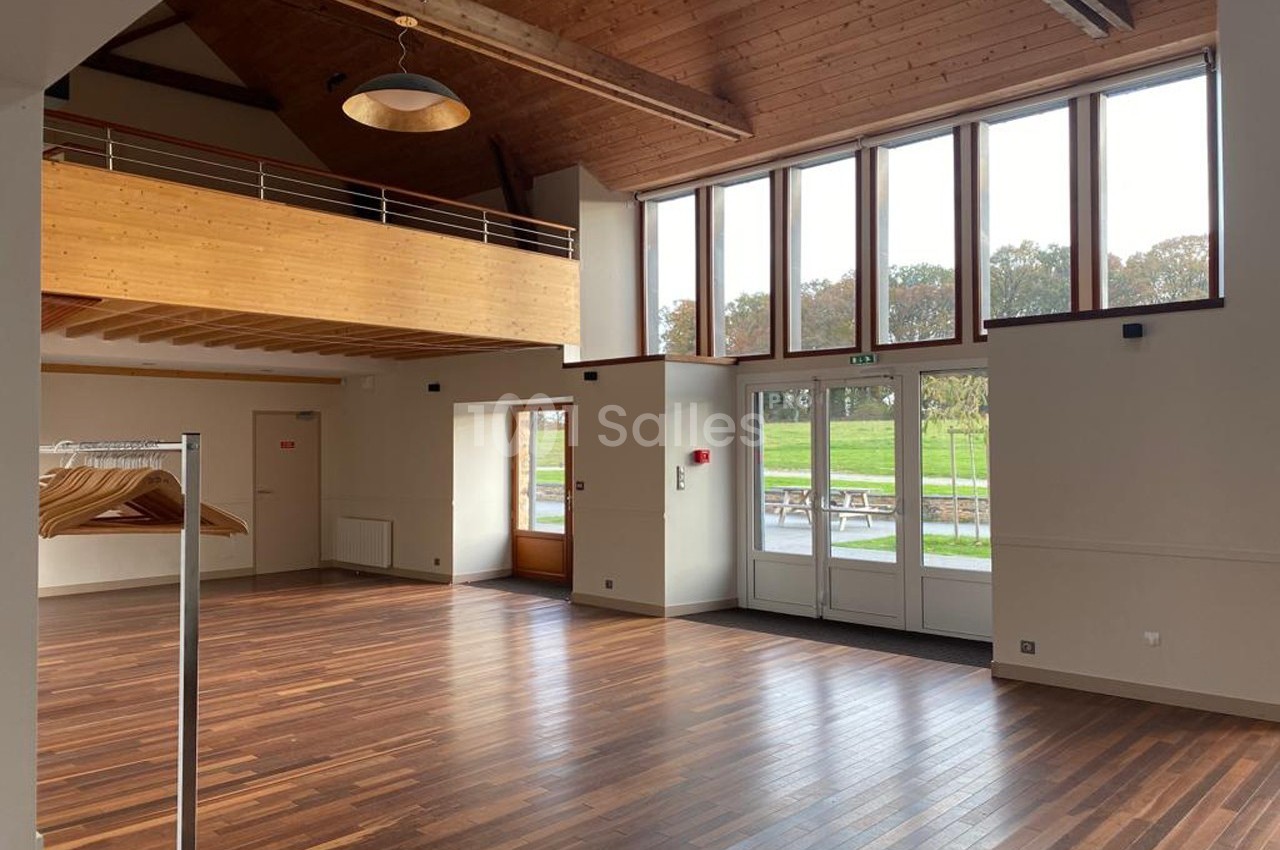 Salle lumineuse avec parquet en bois, grande baie vitrée, mezzanine en bois et vue sur un espace vert extérieur.
