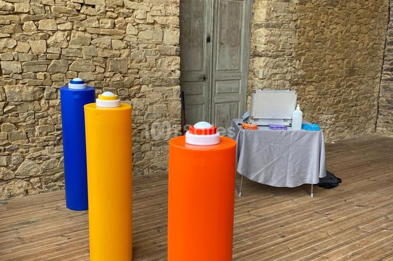 Trois colonnes colorées devant un mur en pierre, avec une table contenant divers objets en arrière-plan.