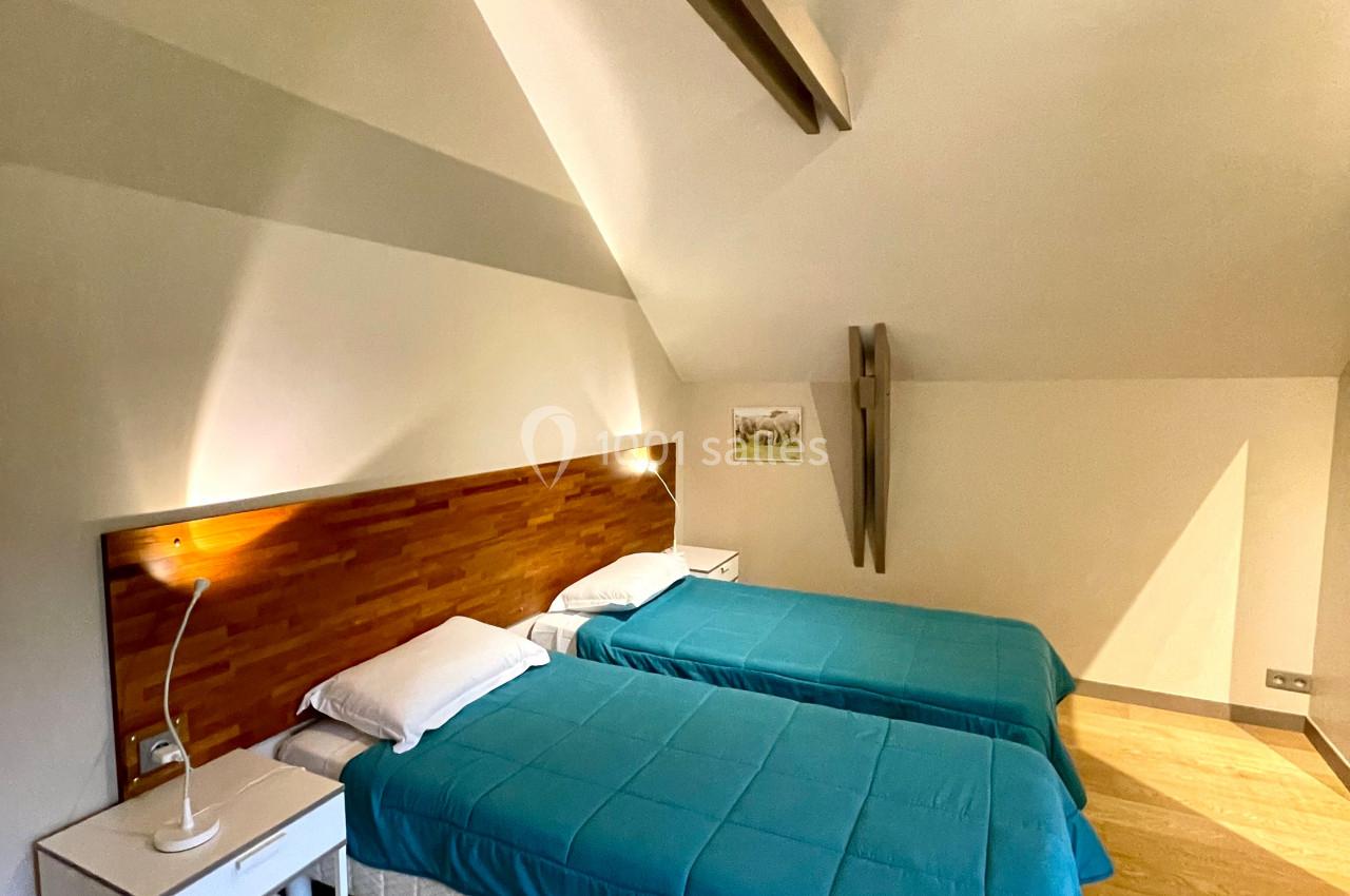 Deux lits simples avec couvre-lits bleus dans une chambre mansardée lumineuse, équipée d'une table de chevet et d'une lampe.