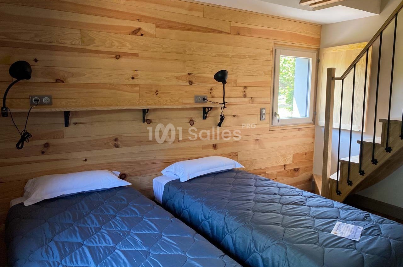 Deux lits simples avec couvre-lits gris dans une chambre aux murs en bois, éclairée par une fenêtre et un escalier visible.
