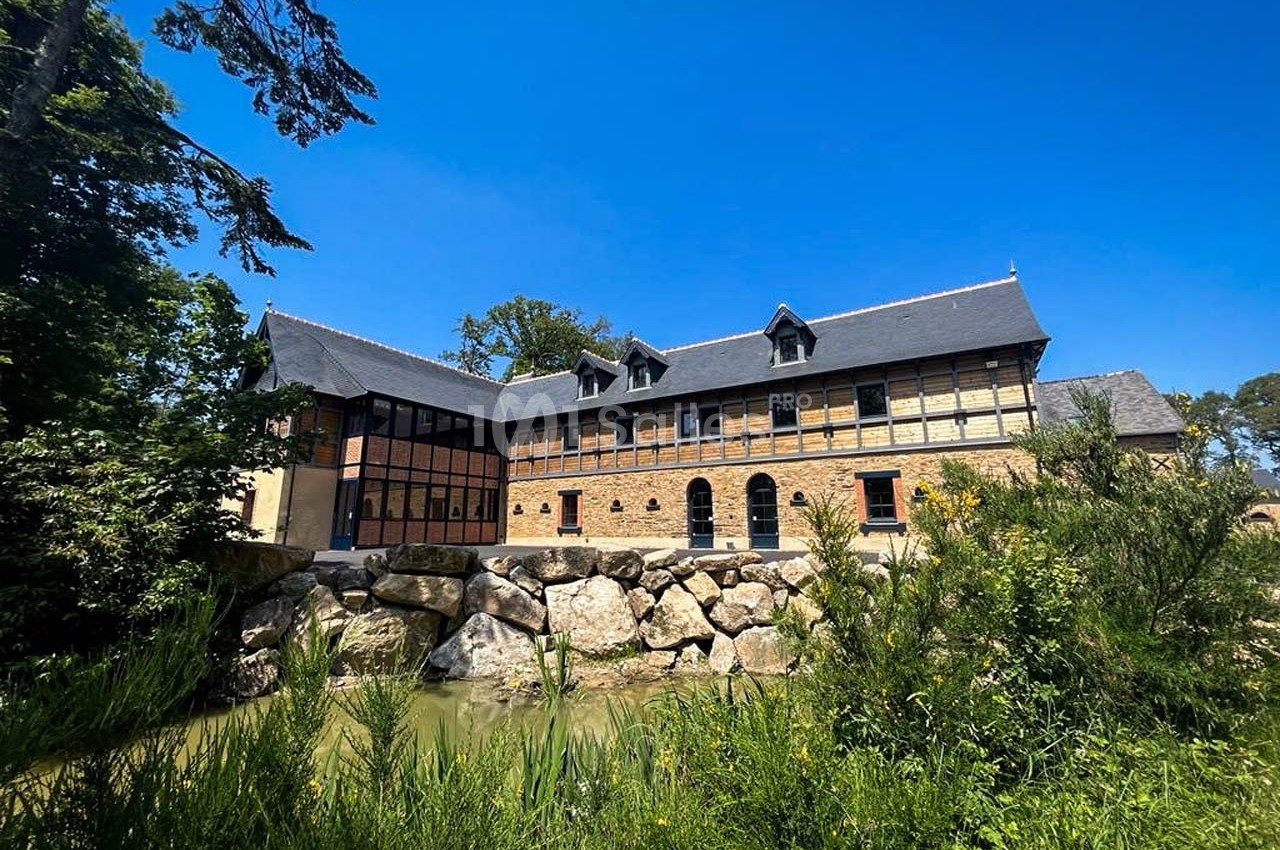 Bâtiment en briques et verre entouré de végétation, avec un ruisseau et des rochers au premier plan sous un ciel bleu.