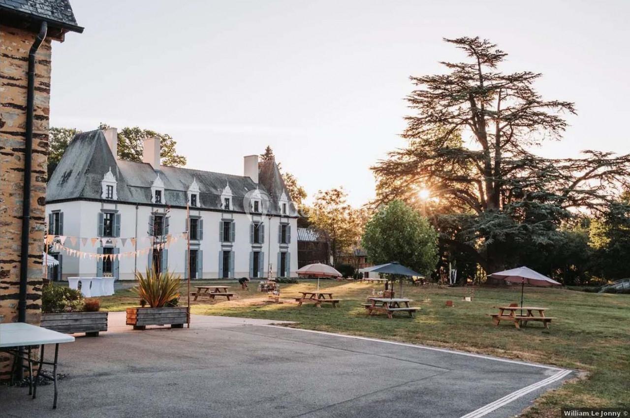 Cour d’un château avec pelouse, tables de pique-nique, parasols et arbres, éclairée par une lumière de fin de journée.