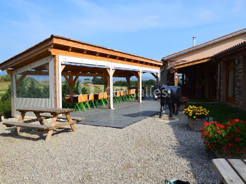 Terrasse couverte en bois avec tables et chaises, entourée de fleurs, d'une statue de vache et d'un paysage rural.