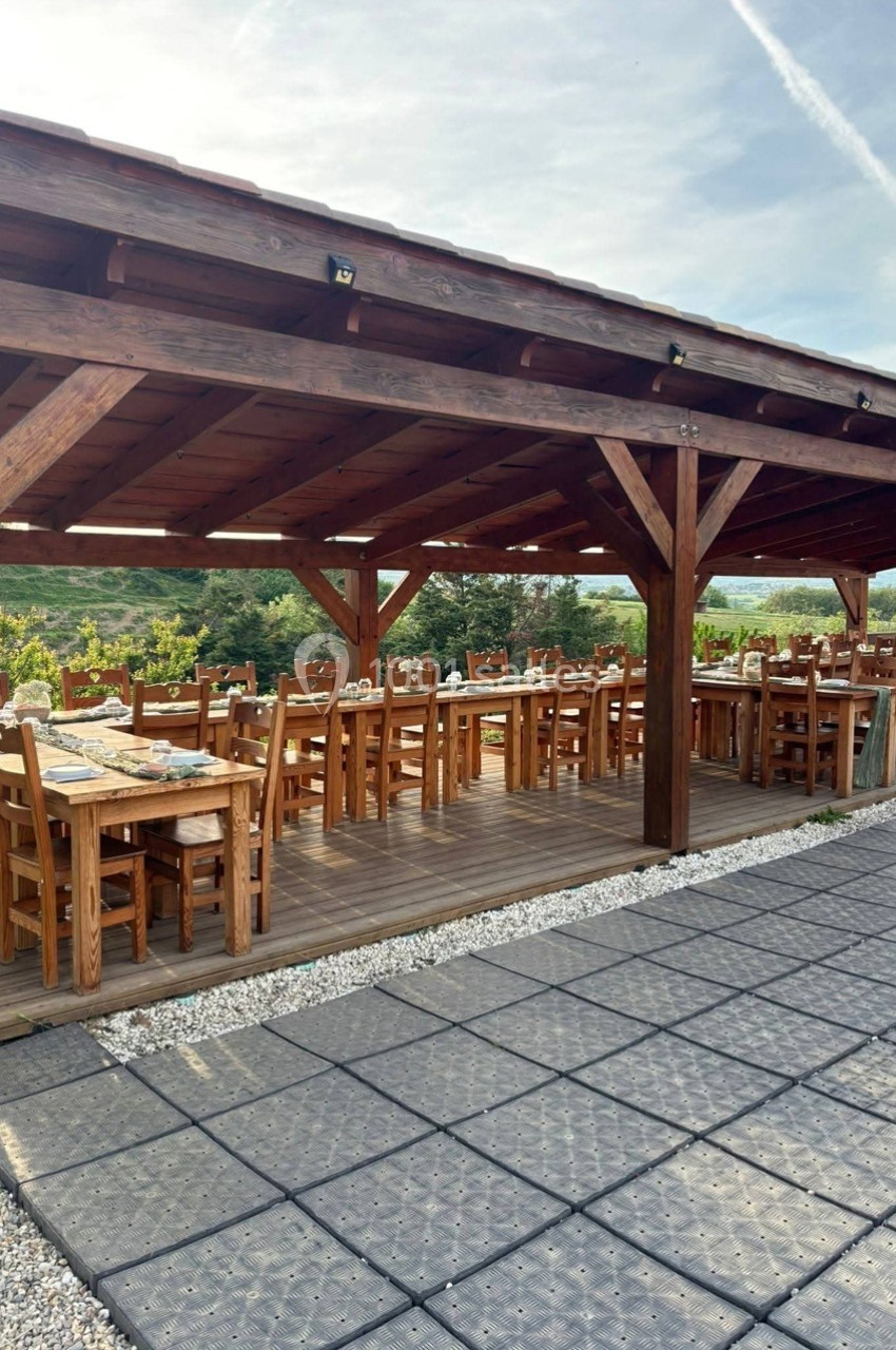 Terrasse en bois couverte avec tables et chaises alignées, entourée de verdure sous un ciel dégagé.