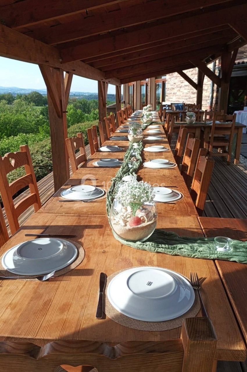 Table en bois dressée pour un repas, décorée de plantes et vases, sous une pergola avec vue sur un paysage verdoyant.