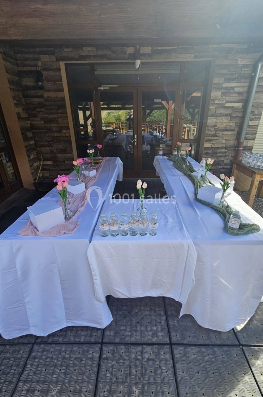 Tables décorées avec nappes blanches, fleurs roses et bouteilles d'eau, disposées en extérieur devant un bâtiment en pierre.