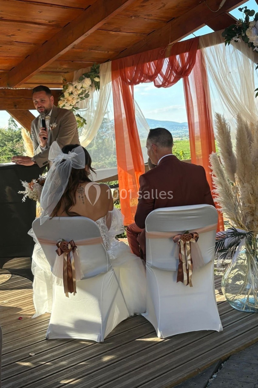 Un couple assis lors d'une cérémonie de mariage en extérieur, avec un officiant s'exprimant sous une pergola décorée.