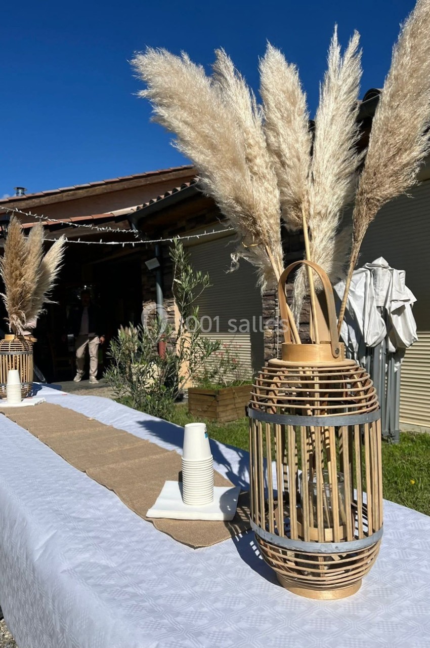 Centre de table avec pampas dans un vase en osier sur une table blanche dressée en extérieur sous un ciel bleu.