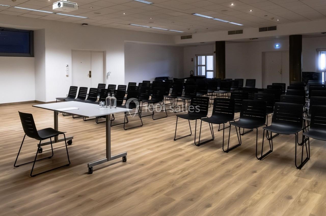 Salle de conférence vide avec des rangées de chaises noires, une table et des bouteilles d'eau sur un sol en bois clair.