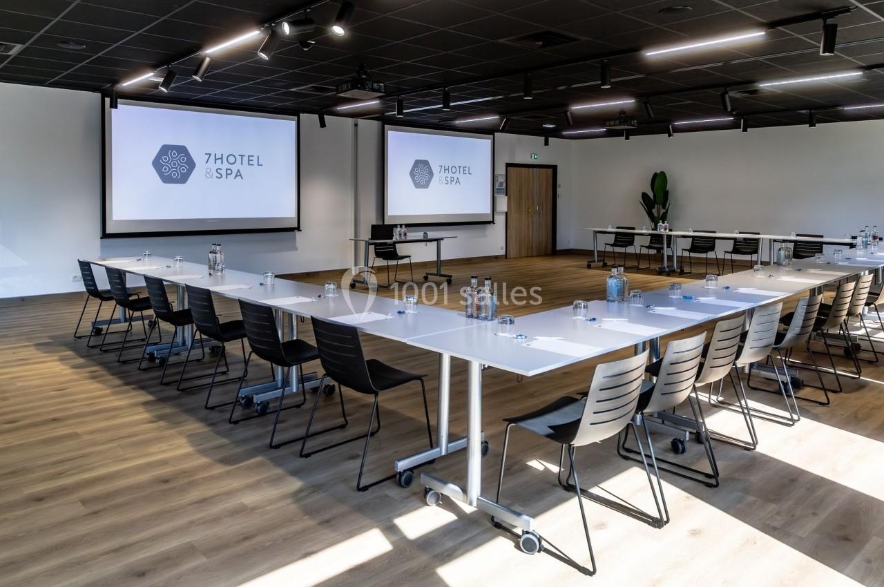 Salle de réunion lumineuse avec tables disposées en U, chaises noires et écrans de projection allumés.