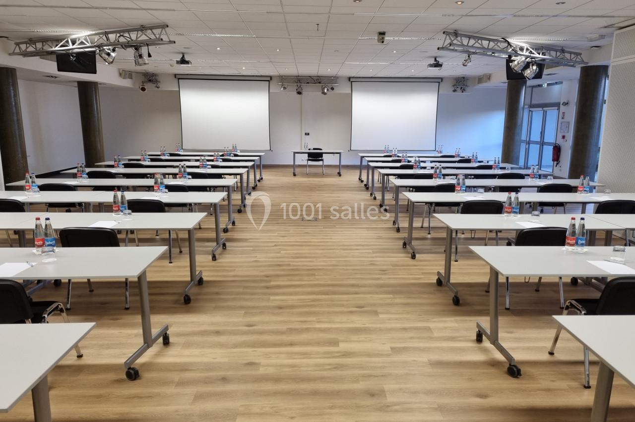 Salle de conférence avec des rangées de tables et chaises alignées, deux écrans de projection et bouteilles d'eau sur les…