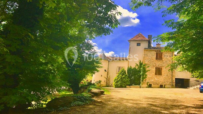 Maison en pierre entourée d'arbres et de verdure, sous un ciel bleu avec quelques nuages.