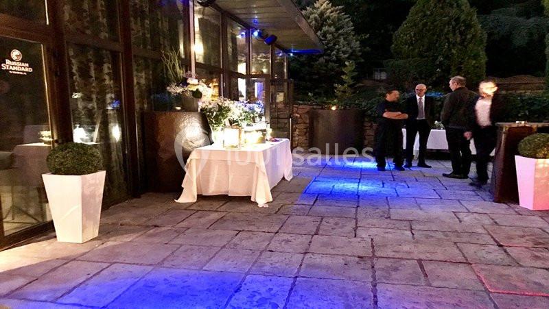 Terrasse éclairée en soirée avec tables décorées, plantes en pots et quelques personnes debout en arrière-plan.