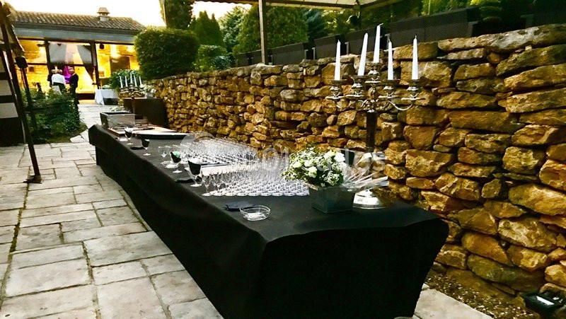 Table dressée en extérieur avec nappes noires, verres, chandeliers et décorations florales devant un mur en pierre.