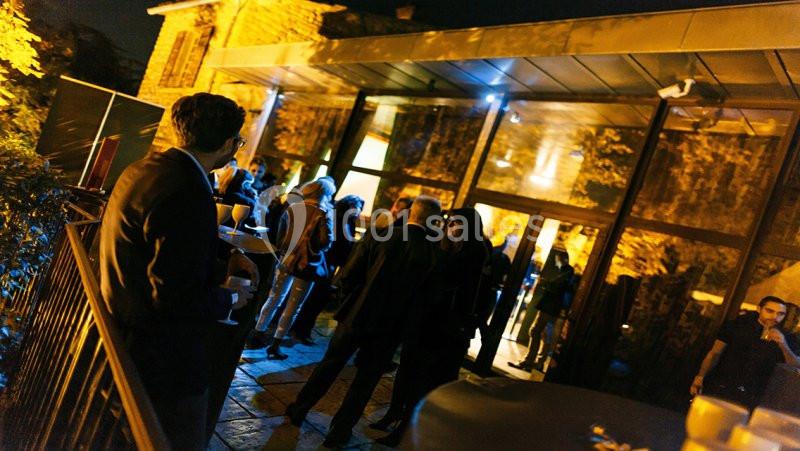 Des personnes discutent et prennent un verre en soirée devant un bâtiment éclairé avec des baies vitrées.