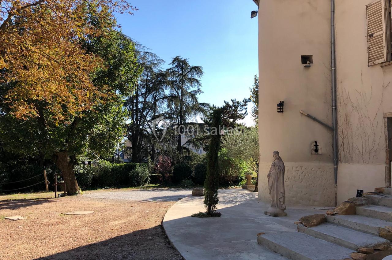 Cour ensoleillée avec statue, façade beige, arbre aux feuilles automnales et végétation en arrière-plan.