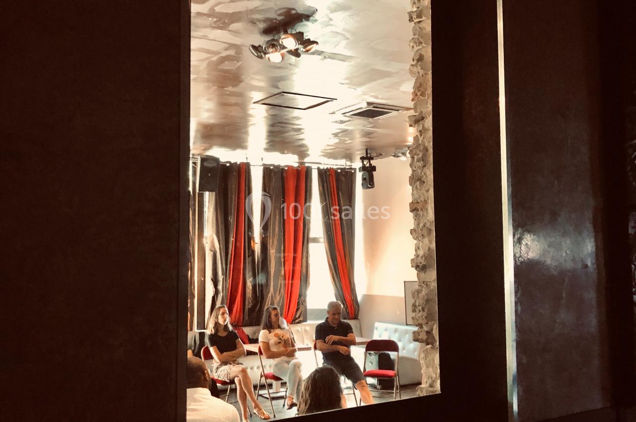 Vue à travers une fenêtre sur un groupe de personnes assises autour de tables dans une pièce aux rideaux rouges et noirs.