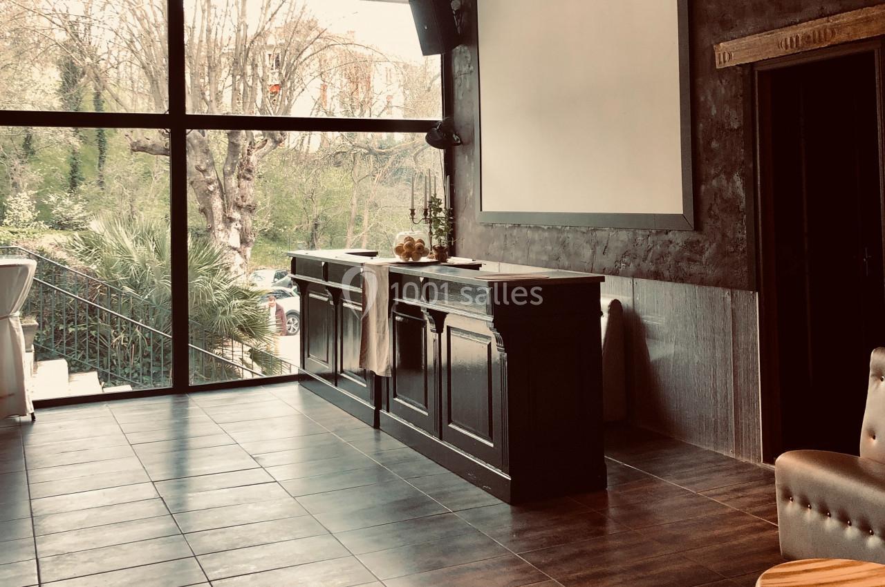Salle lumineuse avec parquet sombre, grand écran blanc, bar en bois et vue sur des arbres à travers une baie vitrée.