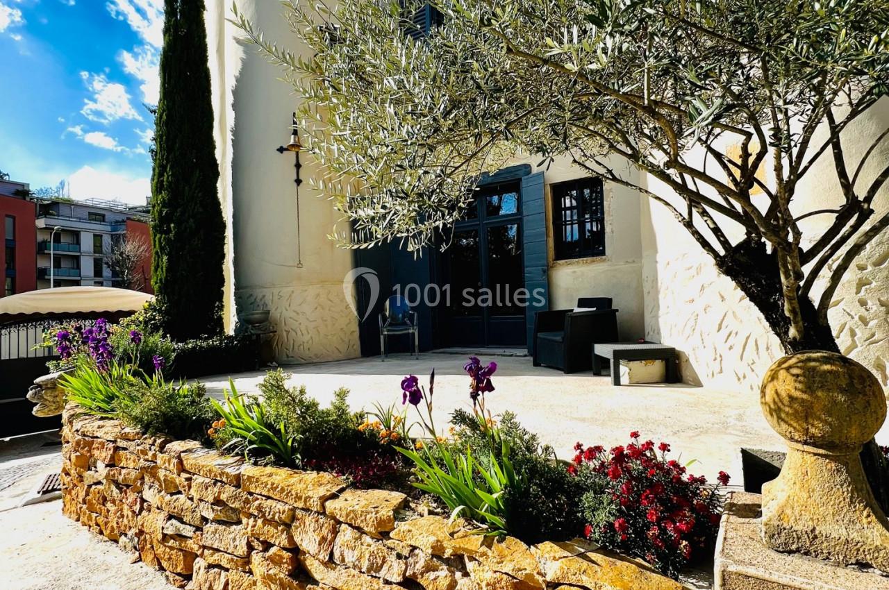 Façade d'un bâtiment blanc avec un olivier, des fleurs colorées et un muret en pierre sous un ciel ensoleillé.
