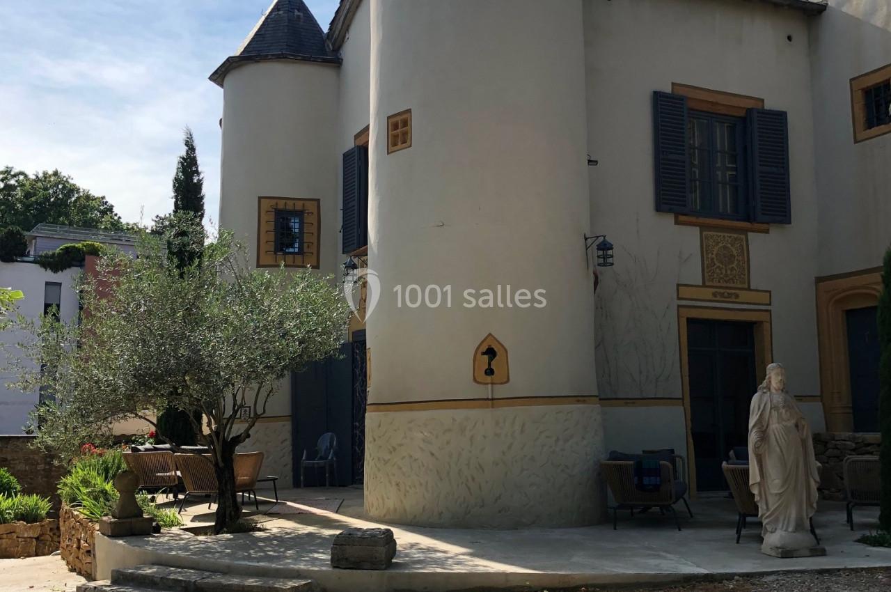 Façade d'une maison avec tours rondes, volets bleus, statue et terrasse dans une cour ensoleillée.