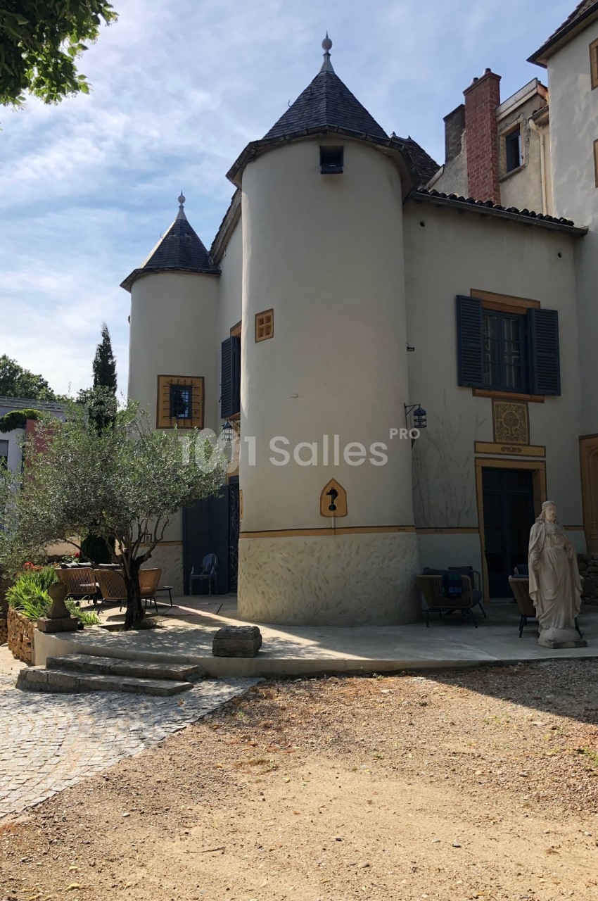 Façade d'une maison avec tours rondes, volets bleus, statue et terrasse dans une cour ensoleillée.