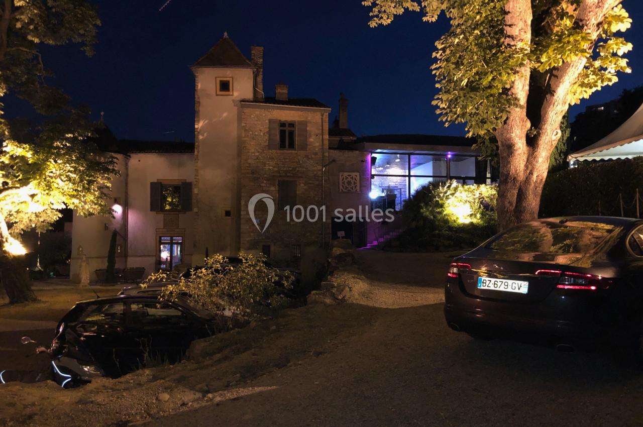 Vue nocturne d'un bâtiment ancien éclairé, entouré d'arbres, avec des voitures garées au premier plan.