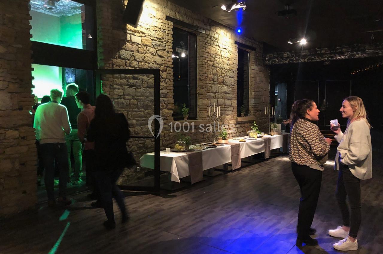 Des personnes discutent dans une salle avec murs en pierre, tandis qu’un groupe se dirige vers une zone éclairée en vert.