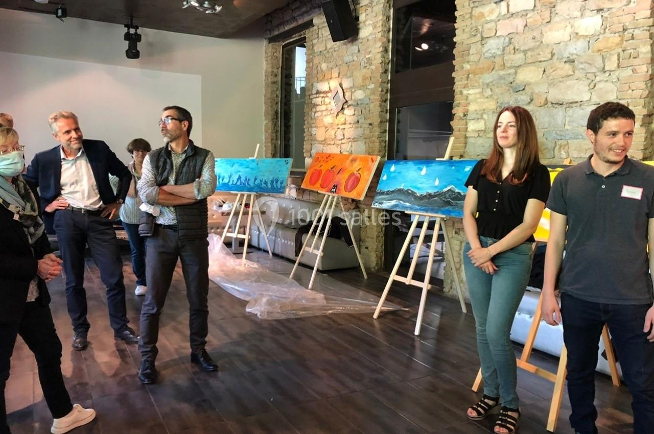 Des personnes debout dans une salle d'exposition avec des peintures colorées sur des chevalets devant un mur en pierre.
