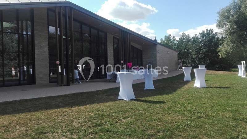 Extérieur d'un bâtiment moderne avec de grandes baies vitrées, tables hautes couvertes de nappes blanches sur une pelouse.