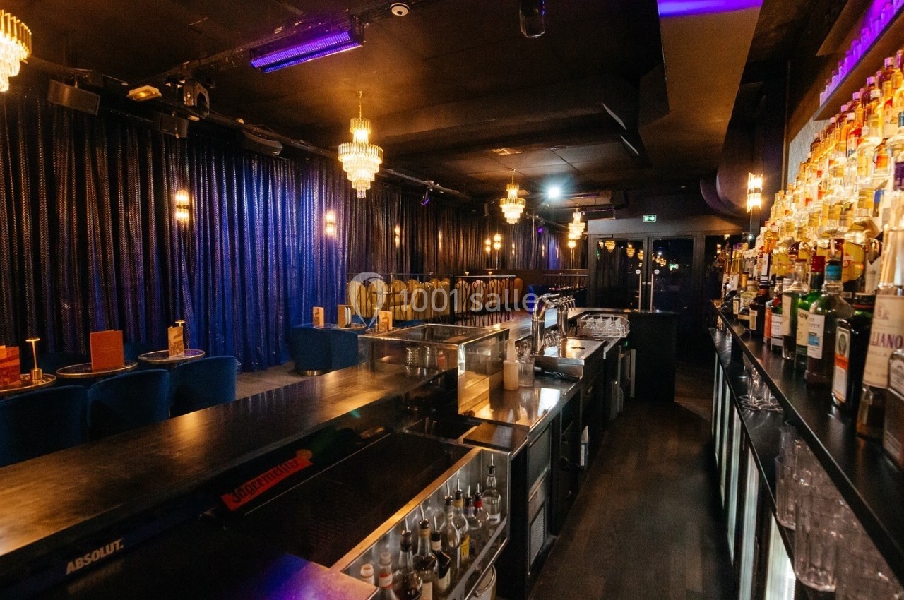 Salle de bar avec comptoir en bois, rangées de bouteilles, tables et chaises sous des lumières tamisées.